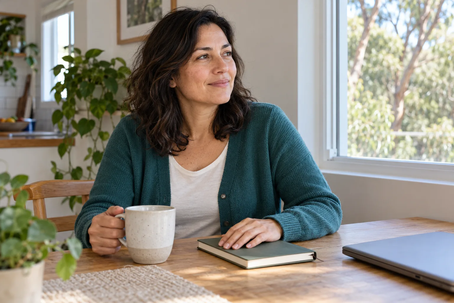 A woman sitting at a sunlit kitchen table with a notebook and mug.